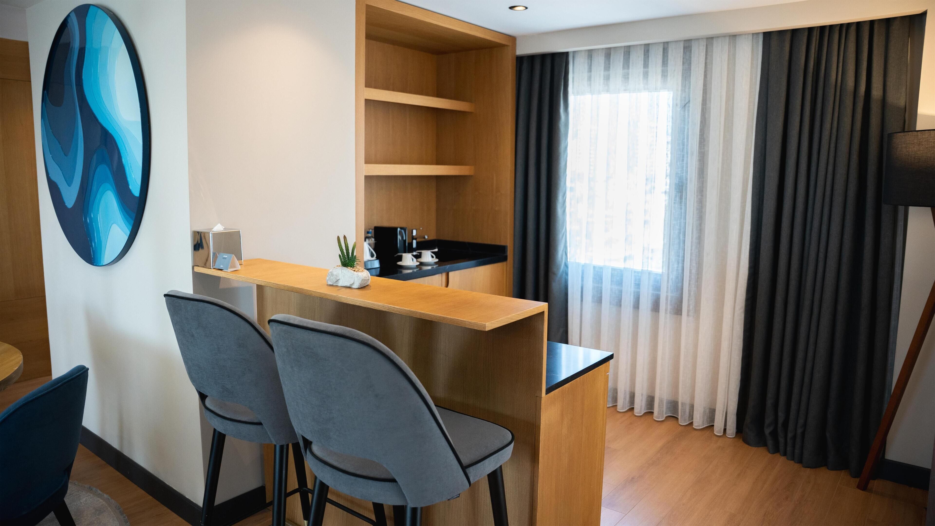 A compact hotel room with a kitchenette, two grey chairs, a wooden desk, a large abstract painting, and a window with sheer curtains.