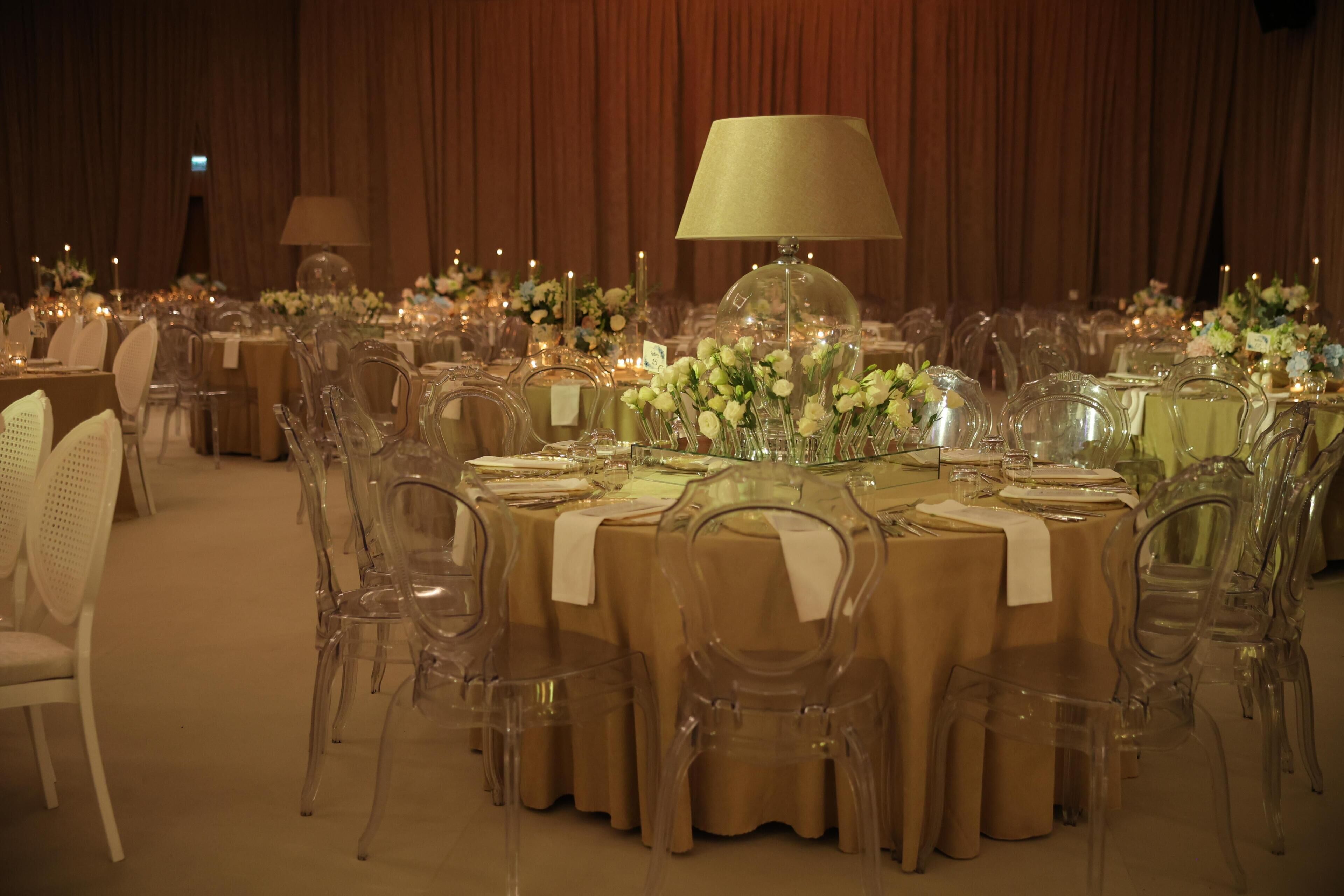 Un salón de banquetes con mesas redondas, sillas traslúcidas y centros de mesa florales.