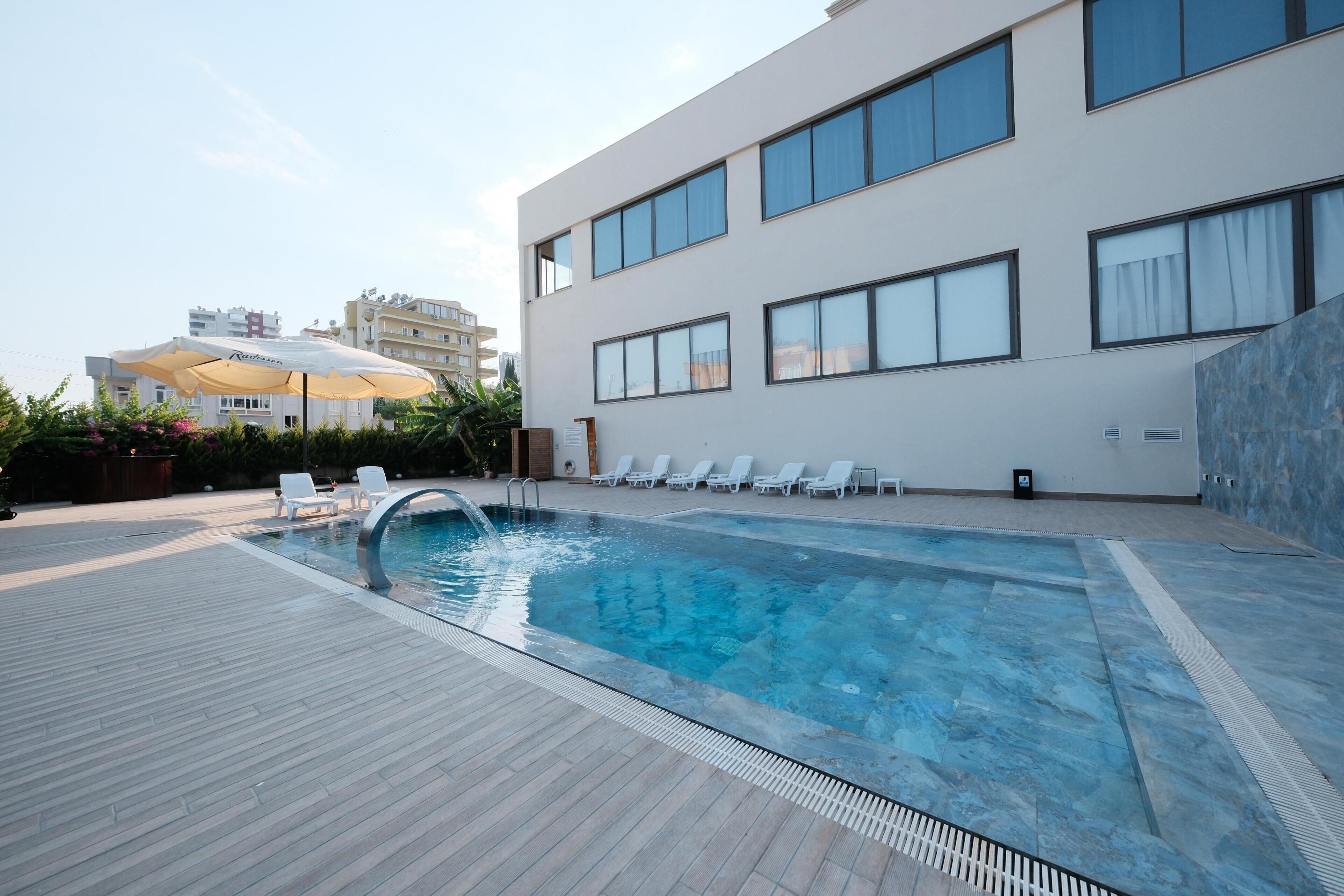 Une piscine avec une terrasse en bois, des chaises longues blanches et un bâtiment moderne aux grandes fenêtres en arrière-plan.