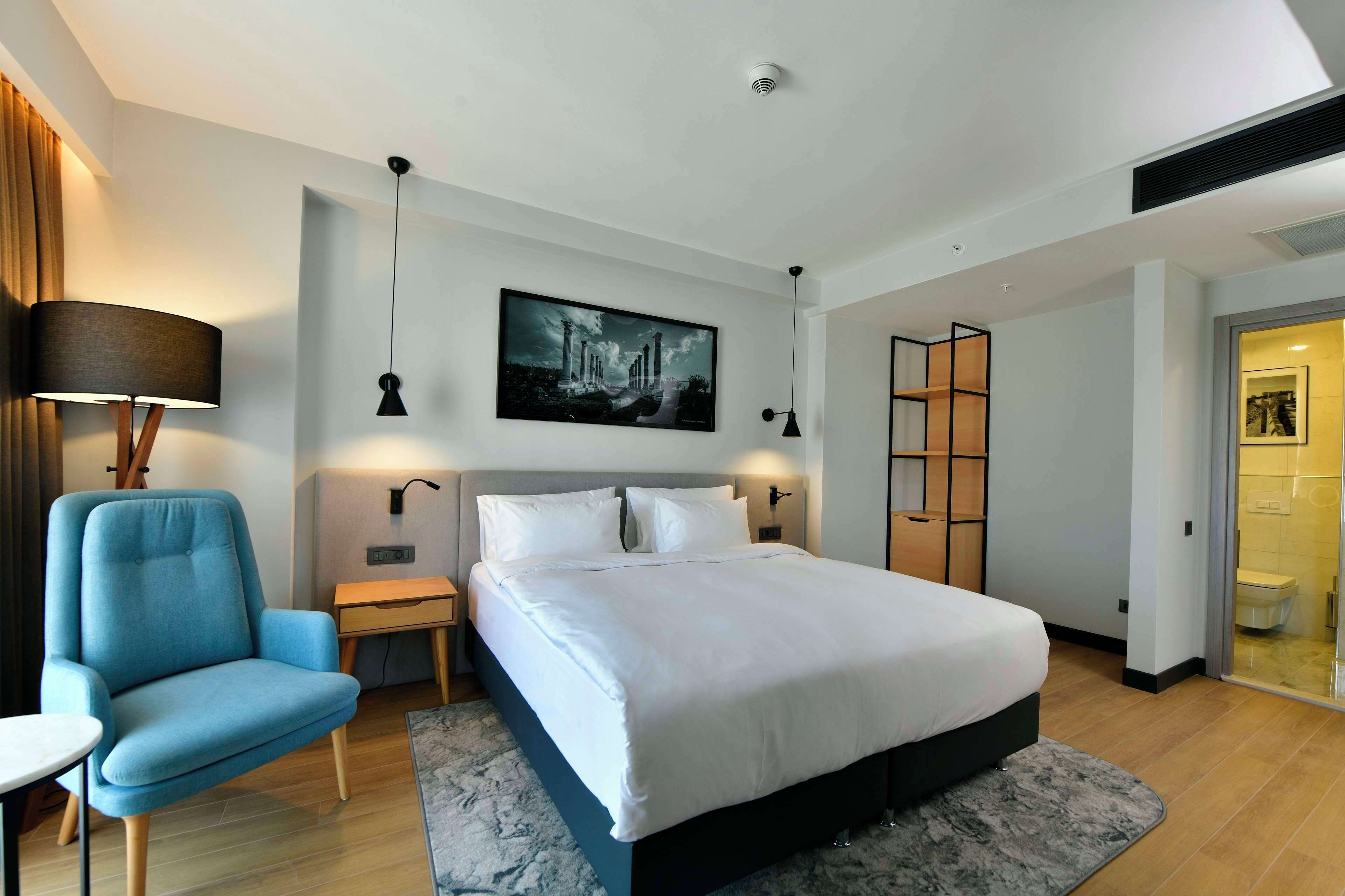 A modern hotel room with a large bed, a blue armchair, a desk, and a painting on the wall.