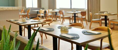 Un vaste espace dédié à la salle à manger, avec de nombreuses tables et chaises, un plafond haut doté d’un éclairage encastré, de grandes fenêtres avec des rideaux et une vue sur le paysage urbain.