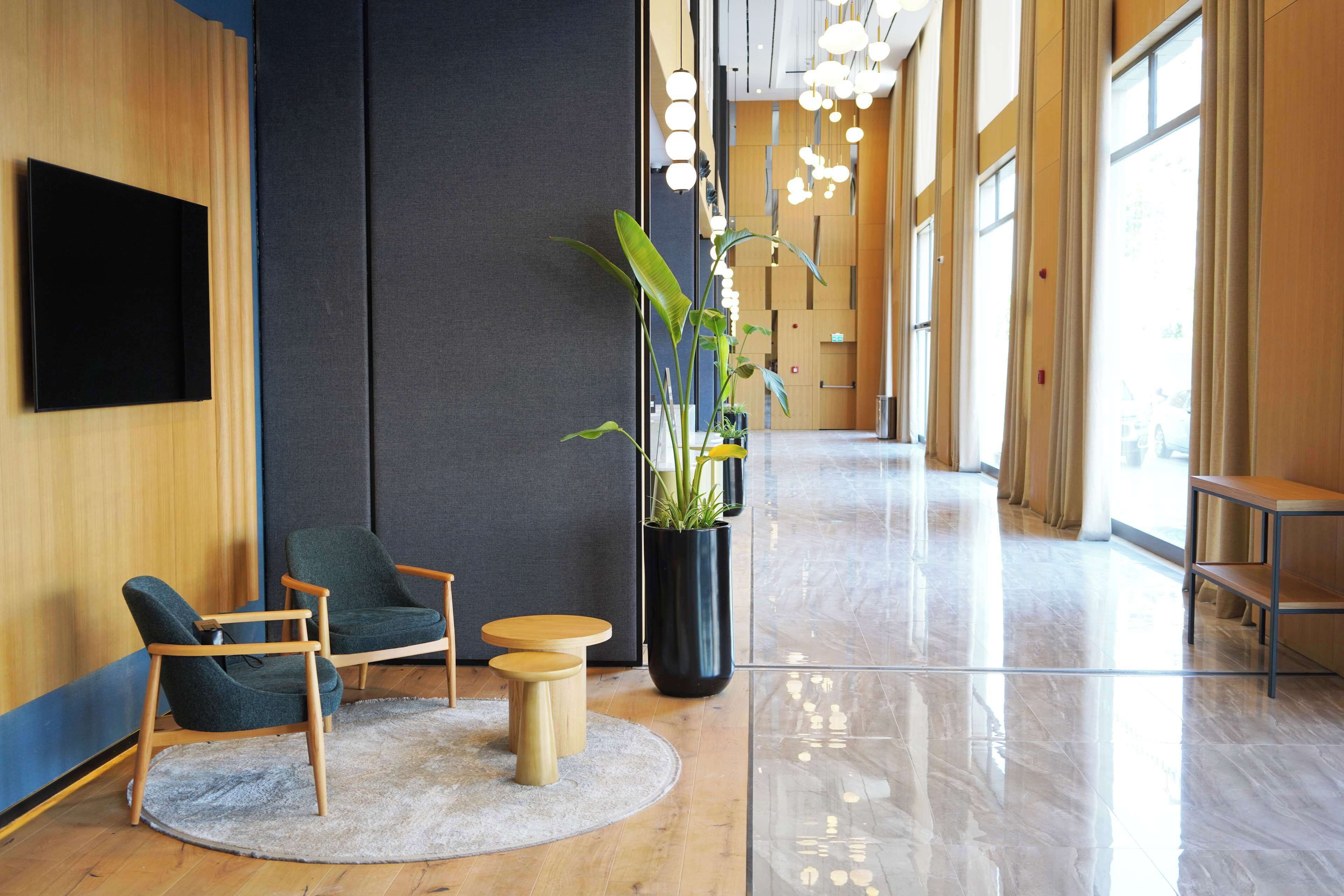 Un vestíbulo moderno con piso de madera, una planta grande en maceta, dos sillas verdes, una mesita redonda y un televisor negro montado en la pared.