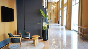 Un vestíbulo moderno con piso de madera, una planta grande en maceta, dos sillas verdes, una mesita redonda y un televisor negro montado en la pared.