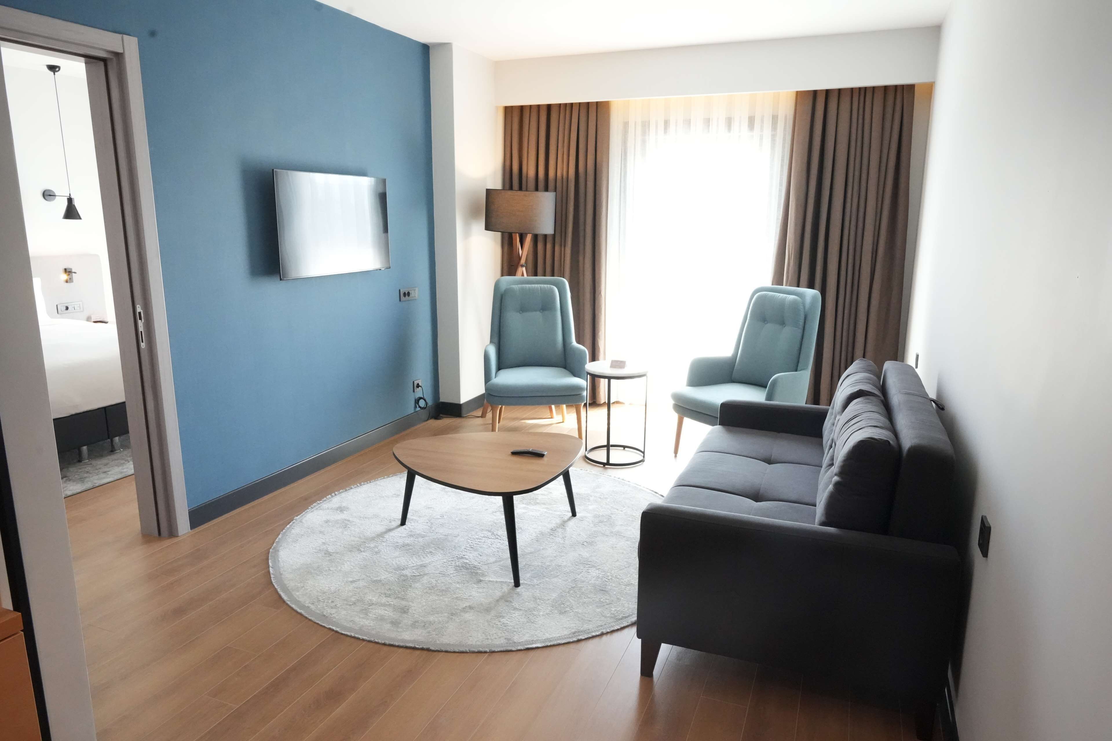 Une chambre d’hôtel moderne avec un mur d’accent bleu, un téléviseur à écran plat, un canapé, deux fauteuils, une table basse ronde et une lampe.