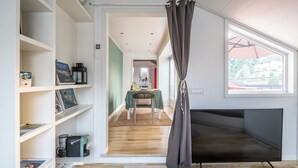 A modern living room with a flat-screen TV, a bookshelf with books and a coffee machine, a dining area with a table and chairs, and a large window with a view of the outdoors. - Lake view attic  (Dagnente)