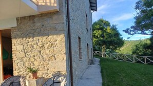 Un bâtiment en pierre avec une terrasse dallée, deux chaises et un petit chien.