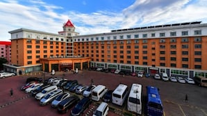 A large hotel with a central entrance, multiple parking spaces, and a red-roofed building in the background. - Friendship Peak Hotel (Burqin)