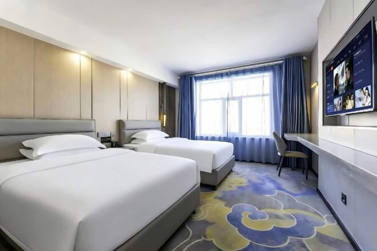 A hotel room with two beds, a desk, a chair, a large window with curtains, and a TV mounted on the wall.