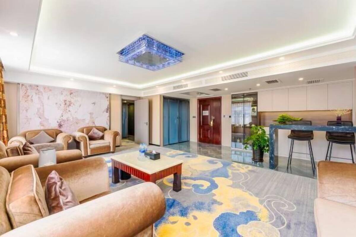A modern hotel lobby with sofas, a coffee table, a patterned rug, a kitchen area, and a ceiling light fixture.