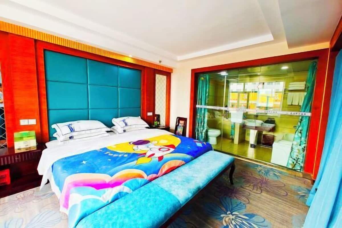 A hotel room with a bed, a bench, a glass partition, and a bathroom visible through the glass.
