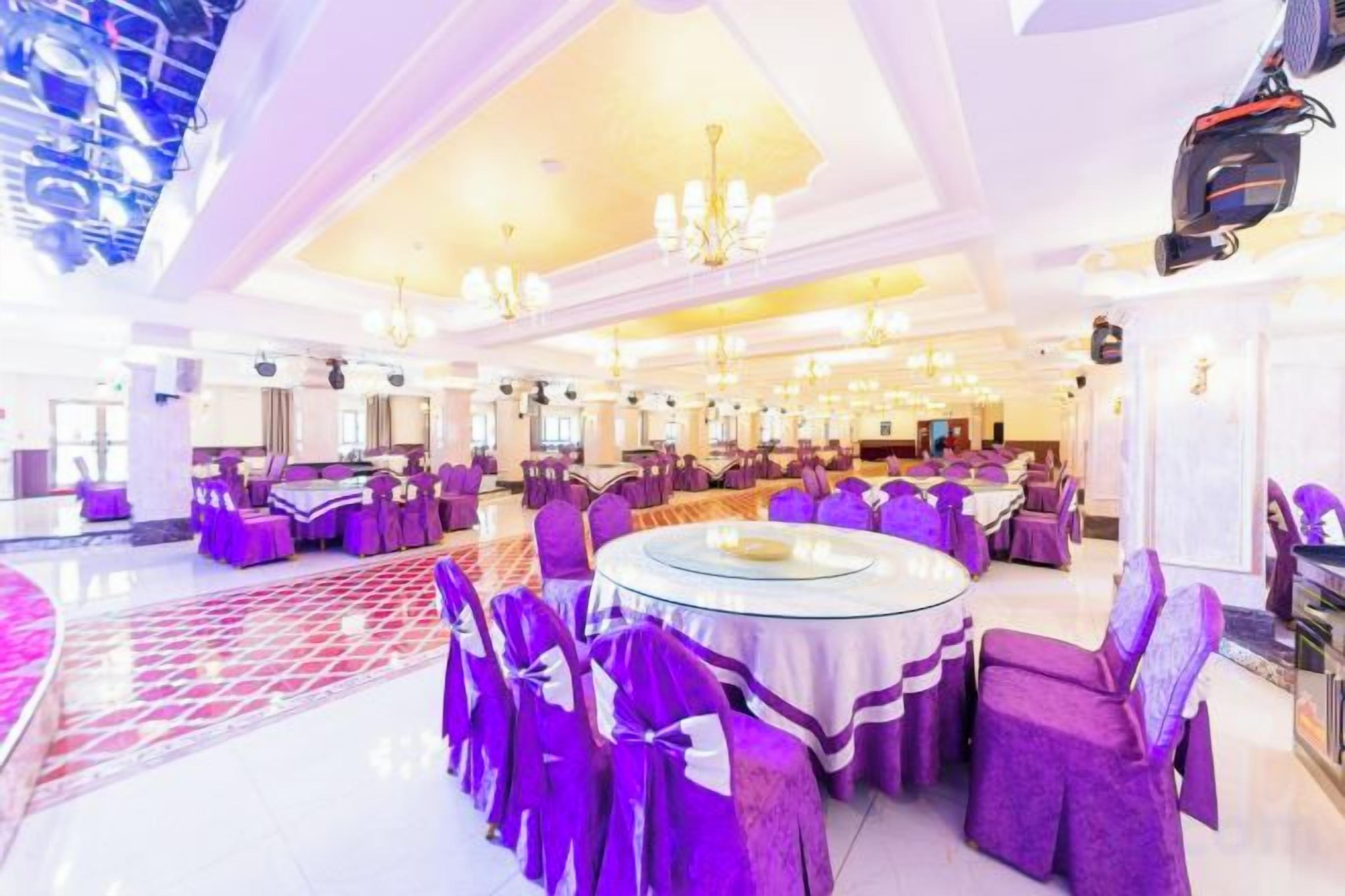Une salle de banquet spacieuse, avec des tables rondes et des chaises violettes, un plafond haut orné de lustres et un plancher ciré.