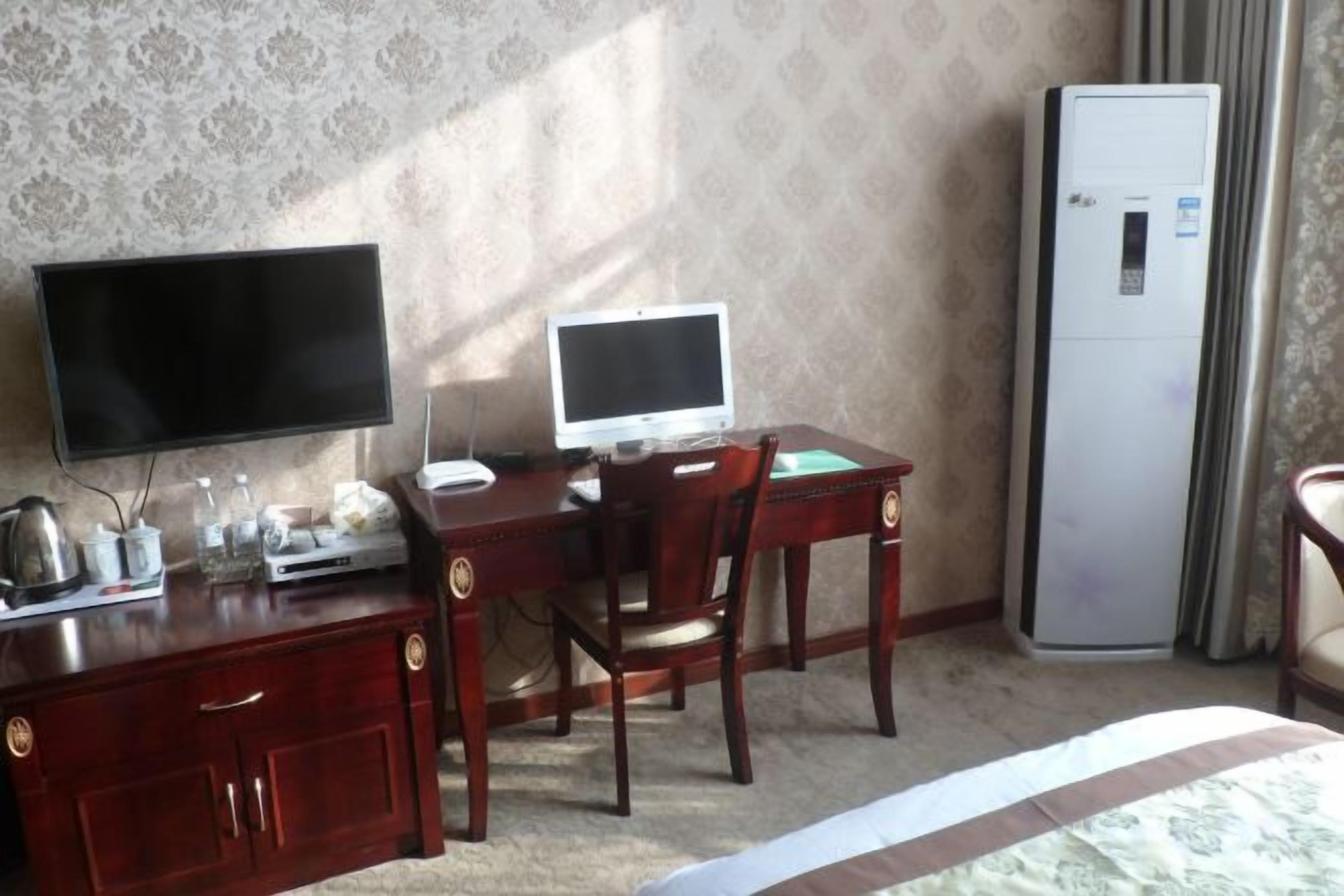 A hotel room with a wooden desk, a television, a computer, a refrigerator, and a bed.