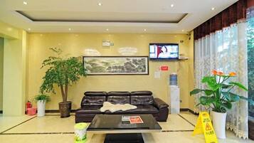 A hotel lobby with a sofa, coffee table, potted plants, a television, and a large window with curtains.