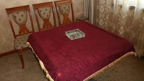 A room with a bed covered in a red blanket, two wooden chairs with patterned cushions, a small table with a glass object, and patterned wallpaper.