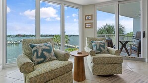 A spacious room with two patterned armchairs, a small wooden table, and large windows offering a view of the water and dock. - Seaspray Riverside 313 (Pensacola)