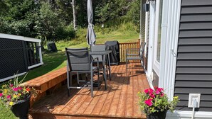 Una terraza de madera con mobiliario exterior, incluyendo sillas y una mesa, rodeada de plantas en macetas.