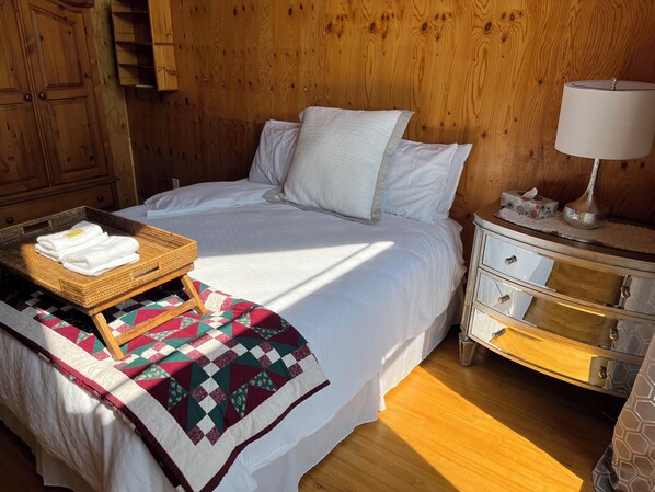 Un lit bien fait, avec du linge de lit blanc, une tête de lit en bois, une table de chevet avec une lampe et un plateau en osier contenant des serviettes pliées.