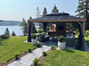 A covered outdoor seating area with a brick fireplace, surrounded by potted plants and outdoor furniture. - Ocean Pearl
Ocean View ,Private Deck W/BBQ, Outdoor Fire Pit (Clarke's Beach)