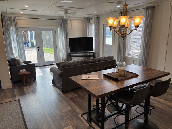 A modern living room with a sofa, a dining table with chairs, a TV, and a chandelier. - Ocean Pearl
Ocean View ,Private Deck W/BBQ, Outdoor Fire Pit (Clarke's Beach)