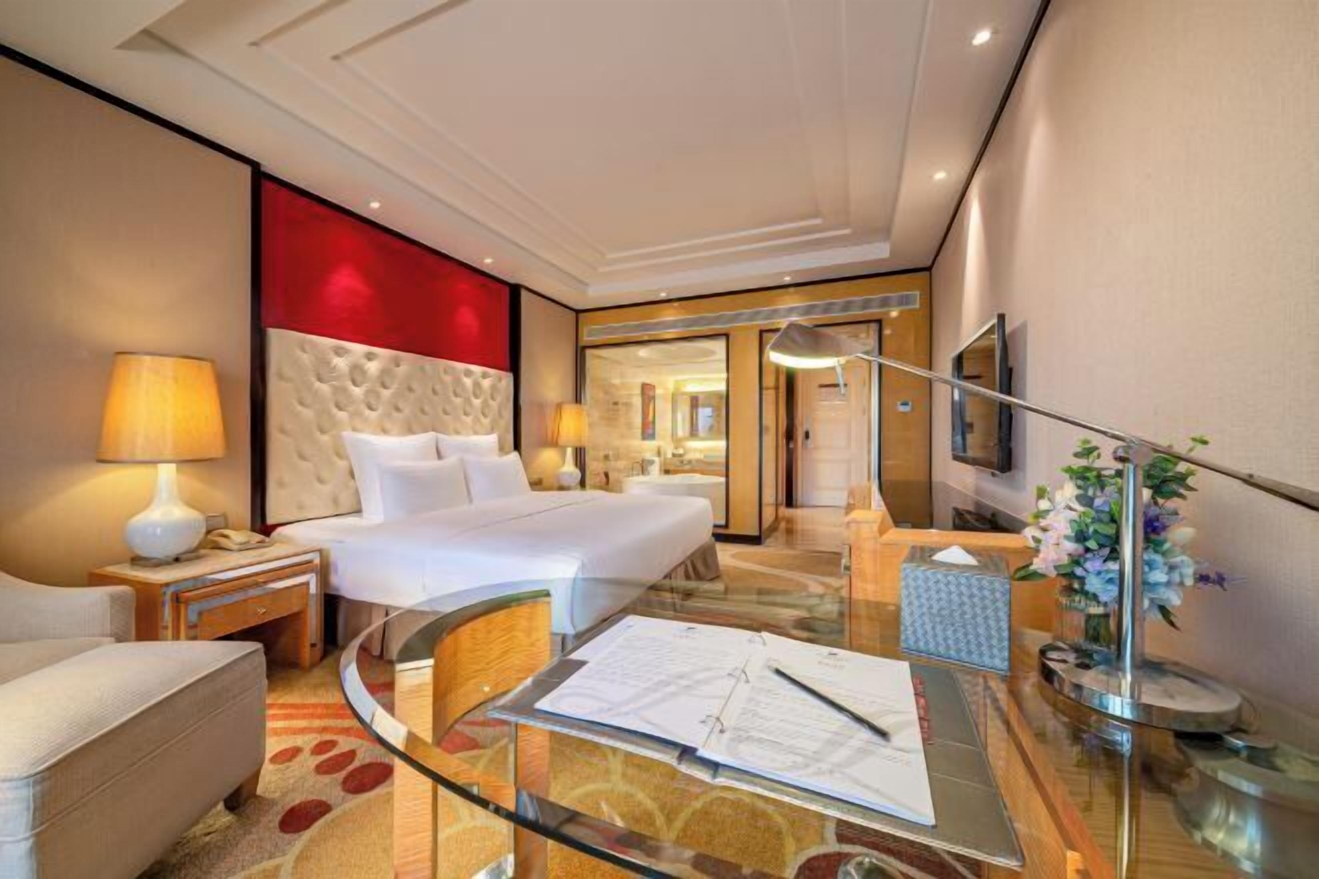 A hotel room with a bed, a desk, a chair, a lamp, a vase with flowers, and a mirror.