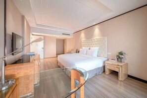 A modern hotel room with a large bed, a glass table, a wooden chair, a bedside table, and a TV.