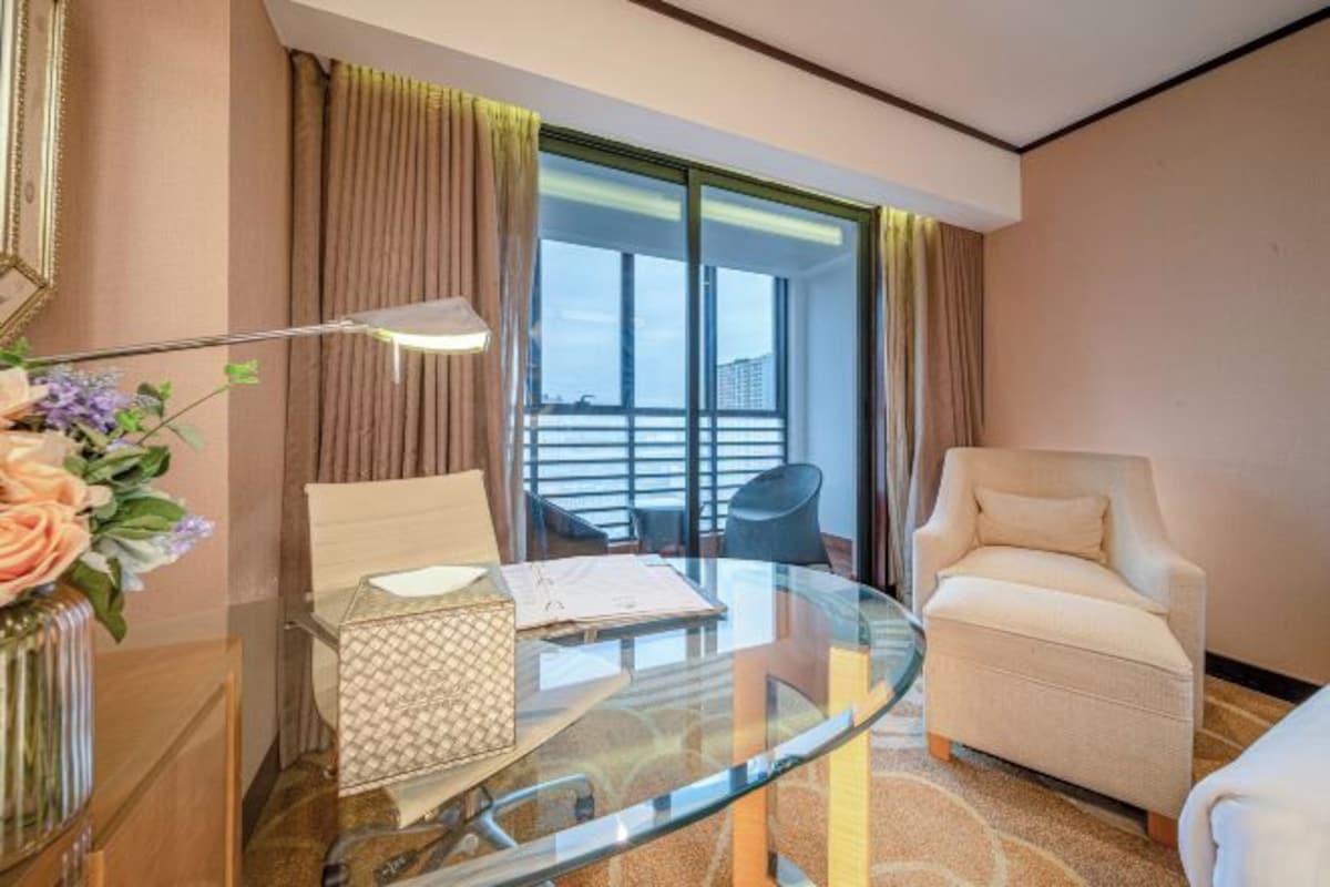 A hotel room with a glass table, a chair, a vase with flowers, a lamp, and a view of the outside.
