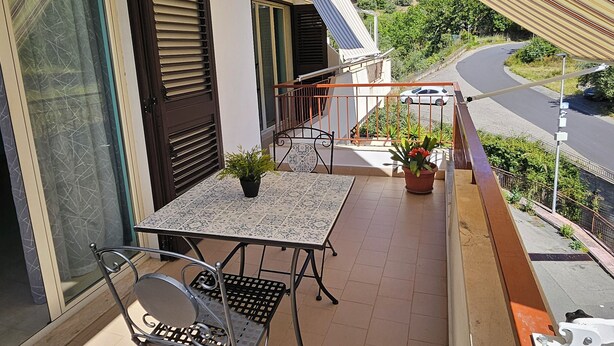 A balcony with a table and chairs, potted plants, and a view of a road and greenery. - Southern Comfort Sicily Holidays (Letojanni)