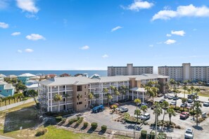A multi-story hotel with a parking lot, surrounded by palm trees and a view of the ocean. - Grand Caribbean East 403 (Pensacola)