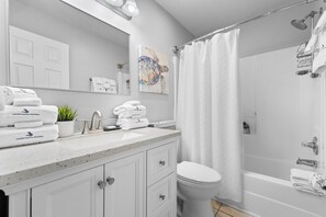 A modern bathroom with a white sink, a toilet, and a shower area. There are towels stacked on the sink and a plant on the counter. - Grand Caribbean East 403 (Pensacola)