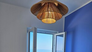 Een kamer met een beige bank, een groot raam met vrij uitzicht op zee en een geweven hanglamp.
