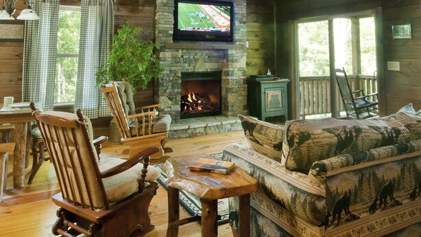 A rustic cabin interior with a stone fireplace, wooden furniture, a television mounted on the wall, and a large window.