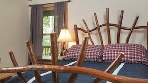 A rustic room with a wooden headboard, a bed with plaid bedding, a window with curtains, and a lamp on a nightstand.