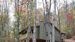 A cabin with a wooden deck and a covered porch, surrounded by trees with autumn foliage.