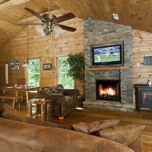 L’intérieur chaleureux d’un chalet avec une cheminée en pierre, un téléviseur fixé au-dessus et un ventilateur de plafond.