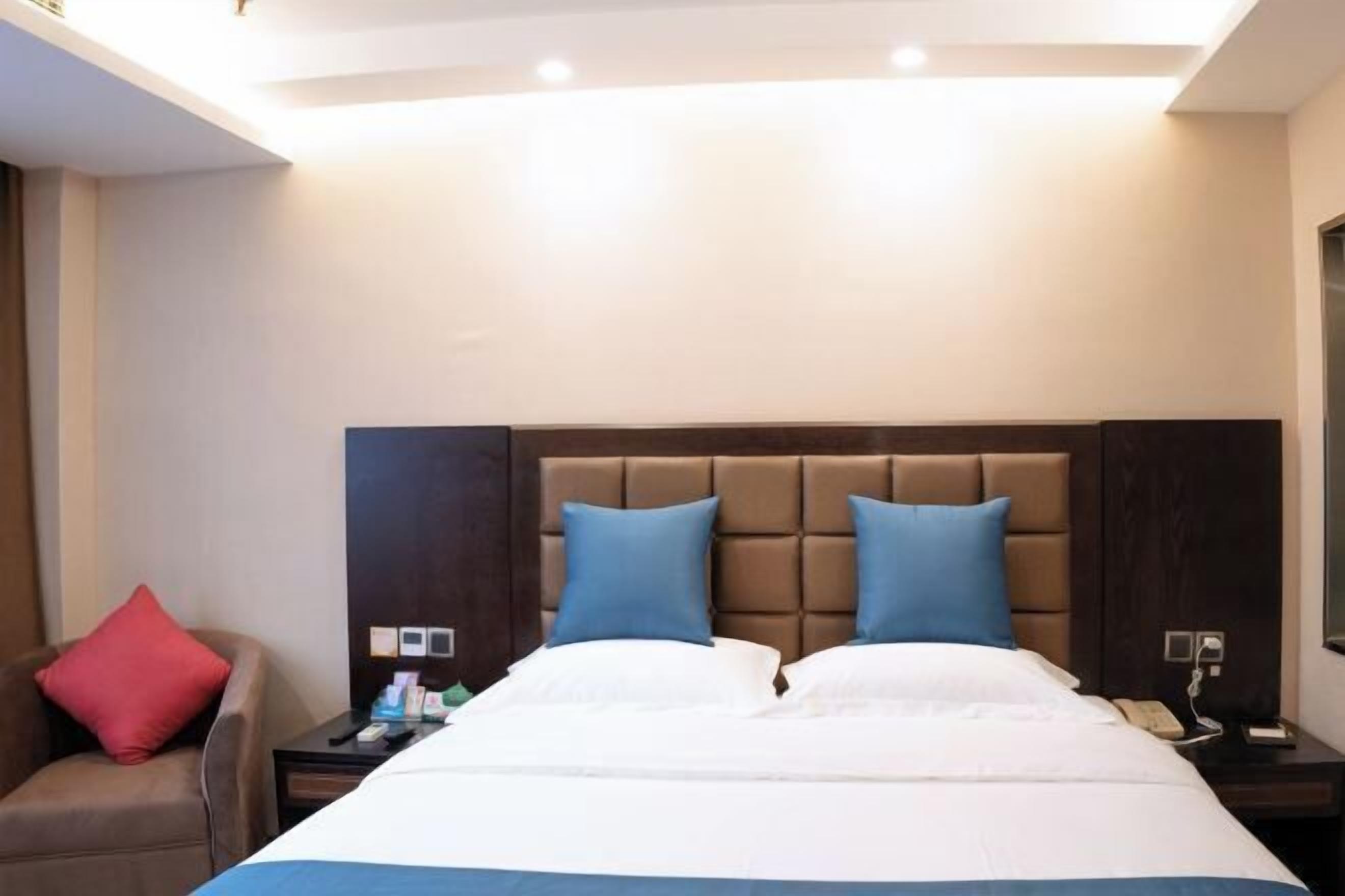 Ein Hotelzimmer mit einem großen Bett, einem braunen Kopfteil, einem roten Kissen, einem Nachttisch mit Telefon und einer Wandleuchte.