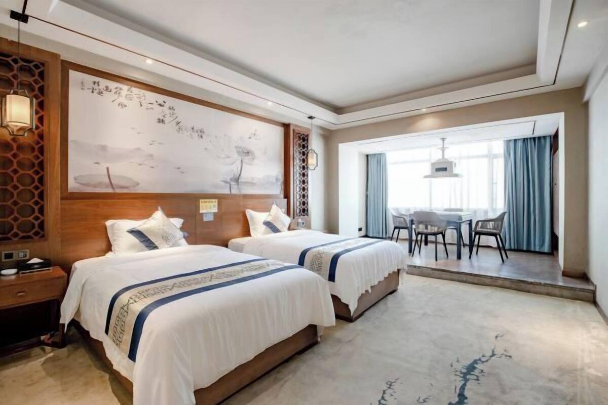 A hotel room with two beds, a large artwork on the wall, a dining area with a table and chairs, and a view through a window.