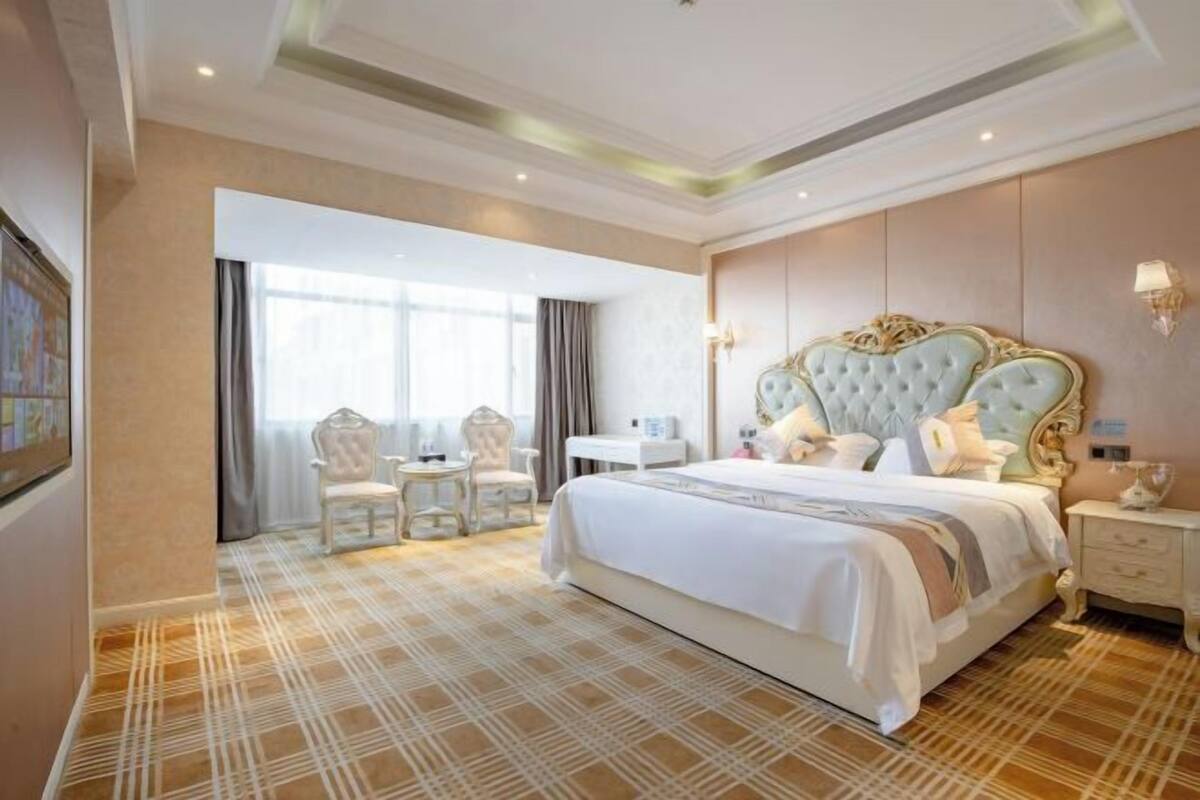 A hotel room with a large bed, a desk, two chairs, and a view through a window.