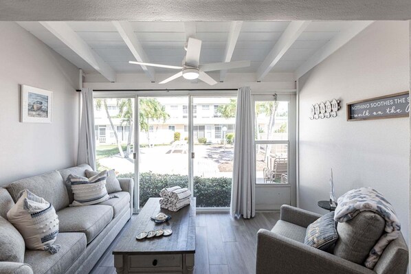A living room with a sofa, a coffee table, and a ceiling fan. There is a view of the outside through large windows.