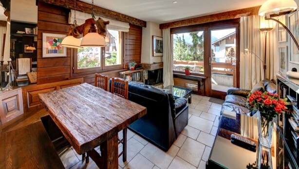 A spacious living area with a wooden dining table, a sofa, and a coffee table. There is a large window providing a view of the outdoors. - Meters from the lift station with a private terrace and balcony in Verbier (Val de Bagnes)