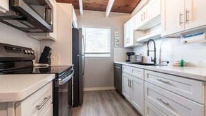 A compact kitchen with a stove, oven, microwave, and refrigerator. There is a window with blinds, and the kitchen has white cabinets and a wooden ceiling.