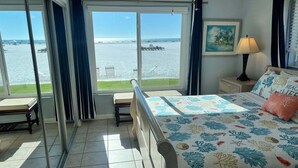 Une chambre à coucher avec un lit, une table de chevet, une lampe, un tableau et une vue sur la mer, visible à travers de grandes fenêtres.