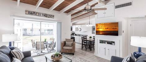 A modern living room with a sofa set, a coffee table, a dining area with chairs, a kitchen with cabinets, a ceiling fan, and a TV displaying a sunset.