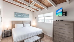 Une chambre à coucher comprenant un lit, une table de chevet, une commode, un ventilateur de plafond, une fenêtre et un mur orné d’un tableau.