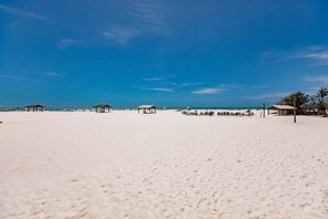 A beach with white sand and clear blue sky. There are several beach huts and lounge chairs.