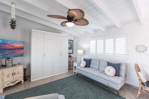 A room with a ceiling fan, a television, a sofa with cushions, a cabinet, and a window with shutters.