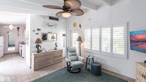 A spacious living room with a ceiling fan, a comfortable armchair, a small table with books, and a large window with blinds.