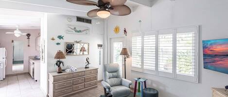 Amplia sala de estar con ventilador de techo, un sillón cómodo, una mesita con libros y un ventanal con persianas.
