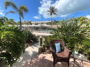 Un'area patio con una sedia in vimini e un tavolino, immersa nel verde e con vista su una piscina.