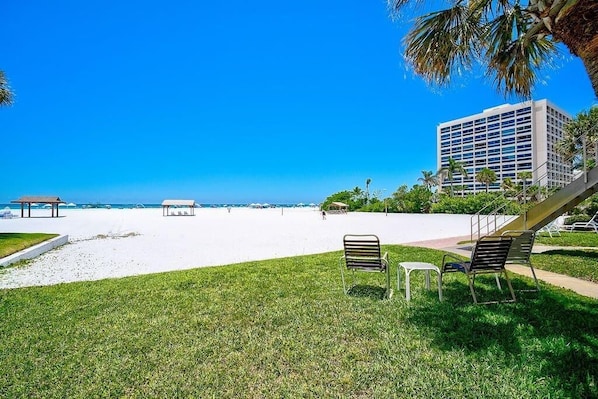 A beach view with a grassy area, a few chairs, and a building in the background.