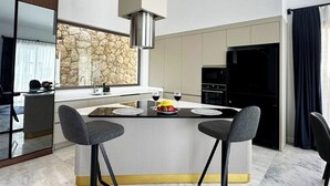 A modern kitchen with a central island, two bar stools, a stone backsplash, and a stainless steel range hood. - Villa Sun 10 - PLANET LUXURY VILLAS (Kas)
